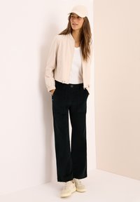 Jongemeisje in een beige pet, lichte beige zipjack, witte shirt, zwarte wijde broek en beige sneakers, staand tegen een lichte muur.