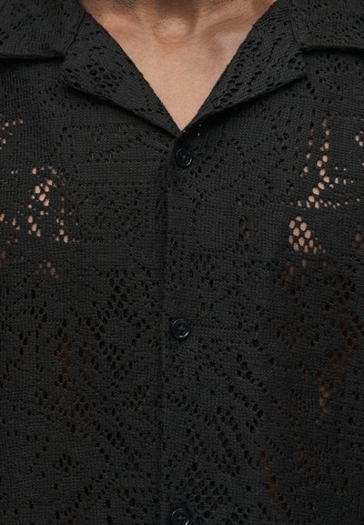 Άνδρας που φοράει μαύρο πλεκτό πουκάμισο με κουμπιά, διακοσμημένο με γεωμετρικά και ανθικά σχέδια, το γιακά ανοιχτός στο λαιμό.