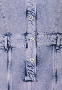 Vaaleansininen farkkukangas, jossa on kolme hopeista nappia, joissa on muurahaislogo, sekä vyölenkki vyötäröalueella.