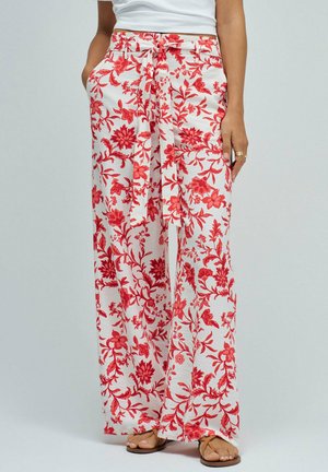 Femme portant un pantalon blanc à jambes larges avec imprimé floral rouge et ceinture en tissu nouée, assorti à des sandales marron clair et un haut blanc.
