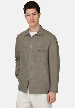 Młody mężczyzna w jasnobrązowej kurtce zapinanej na guziki z dwoma kieszeniami na piersiach oraz w białych spodniach na jednolitym tle.