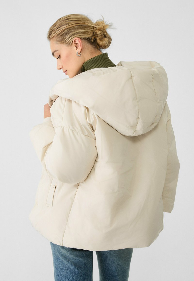 Giacca puffer beige chiaro con cappuccio, caratterizzata da una forma squadrata, texture trapuntata e tasche laterali, indossata sopra un maglione verde scuro a collo alto.
