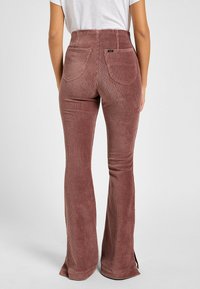 Donna che indossa pantaloni in corda mauve a vita alta e svasati con tasche posteriori e una camicia bianca infilata, in piedi davanti a uno sfondo neutro.