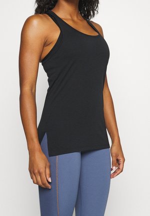 Femme portant un débardeur de sport noir sans manches et un legging bleu, debout devant un fond blanc uni.