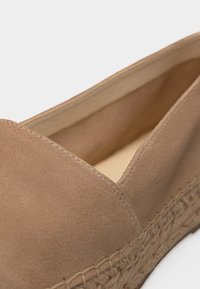 Chaussure en daim marron clair sans lacets avec semelle en jute tressé et coutures visibles le long du bord supérieur et de la semelle.