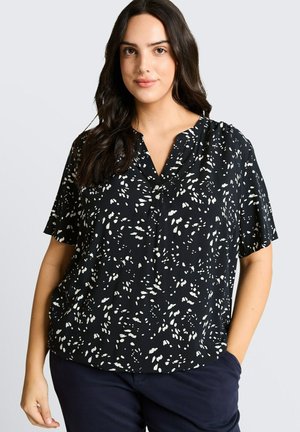 Kobieta ubrana w czarną bluzkę z krótkim rękawem z białymi abstrakcyjnymi plamami i granatowe spodnie, stojąca z jedną ręką w kieszeni na jednolitym tle.