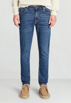 Jean en denim bleu avec une coupe droite, présentant un léger effet délavé, des usures légères et des coutures contrastées, associé à des chaussures décontractées couleur beige.