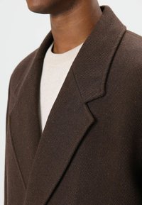 Bruine wolblazer met visgraatmotief, opvallende revers, gestructureerde stof en getailleerde pasvorm, gedragen over een lichtgrijze trui.