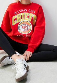 Sudadera roja de los Kansas City Chiefs con texto en blanco y amarillo, que presenta un logo. Combinada con leggings negros y zapatillas blancas con toques en gris.