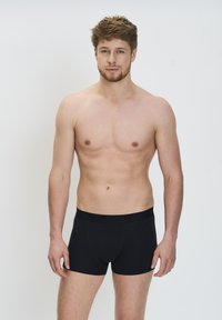 Fiatal férfi világosbarna hajjal és szakállal, fekete, testhezálló boxeralsóban áll egy sima világos háttér előtt.
