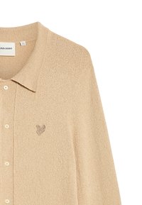 Beige kofta gjord av strukturerad tyg med krage, med knäppning framtill och en liten, broderad logotyp på vänster bröst.