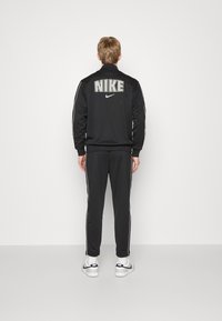 Svart Nike-träningsoverall med vita sidostråk. Baksidan har stora "NIKE"-bokstäver och logotyp. Inkluderar ärmar och byxor med manschetter, används med sneakers.