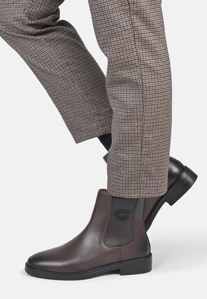 Person wearing brown leather Chelsea boots and checkered slim-fit trousers standing with one foot crossed behind the other.