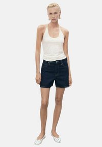Shorts di denim con un lavaggio scuro, taglio a vita media e tasche frontali, abbinati a una canottiera bianca senza maniche con piccole decorazioni.