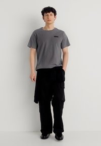 Νεαρός άνδρας που στέκεται δίπλα σε λευκό τοίχο, φορώντας γκρι μπλουζάκι Levi's, μαύρο παντελόνι cargo και μαύρα παπούτσια, με μαύρο μπουφάν δεμένο γύρω από τη μέση.