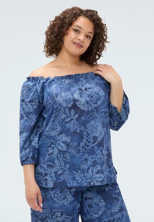 Femme aux cheveux bouclés portant un haut bleu à fleurs épaules dénudées et un pantalon assorti, tenant l'encolure du haut d'une main.