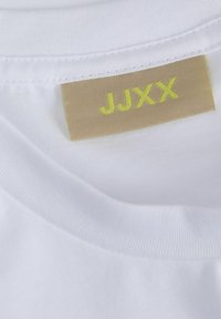 Bílá bavlněná tričko s kulatým výstřihem, s hnědou látkovou nášivkou s nápisem "JJXX" v jasně žlutém písmu.