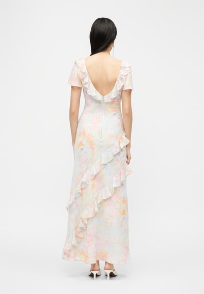 Γυναίκα που φοράει φορεματίνα maxi με παστέλ λουλουδάτο σχέδιο, με βολάν, κοντά μανίκια, βαθιά V στην πλάτη και λευκά σανδάλια με τακούνι, στέκεται γυρισμένη προς τα πίσω.