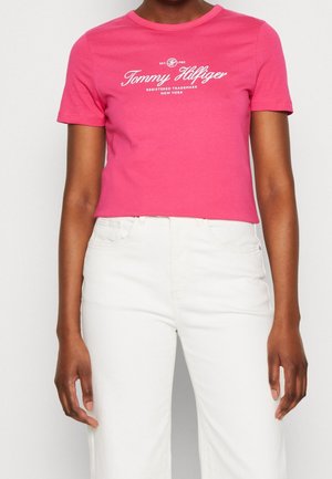 Osoba nosząca różową koszulkę Tommy Hilfiger i białe spodnie z wysokim stanem, stojąca z rękami swobodnie wzdłuż ciała.