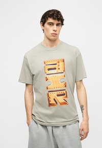 Νεαρός άνδρας με σγουρά μαλλιά, φοράει γκρι μπλουζάκι με μεγάλο πορτοκαλί 3D κείμενο και γκρι φόρμα, στέκεται με το ένα χέρι στην τσέπη.