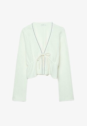 Cardigan court blanc à manches longues avec col en V, petits clous noirs le long de l'encolure et fermeture à nouer à l'avant à la taille.