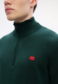 Dunkelgrüner Strickpullover mit einem Halbreißverschluss am Halsausschnitt, ausgestattet mit einem strukturierten Rippkragen und einem kleinen roten "HUGO"-Logopatch auf der Brust.