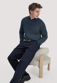 Donker teal gebreid sweater met een ronde hals, gecombineerd met marineblauwe broek en zwarte schoenen, zittend op een lichtgekleurde gestoffeerde kruk.