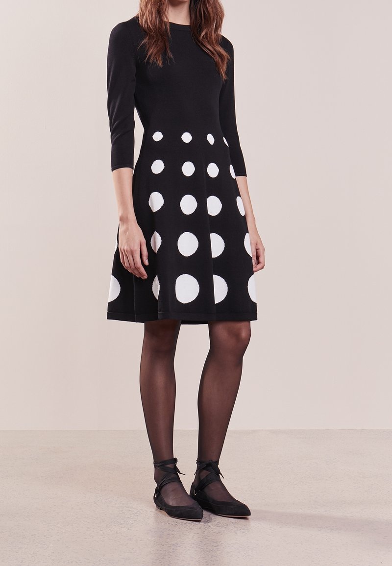 Femme portant une robe noire mi-longue à pois blancs, manches trois-quarts, collants noirs transparents, et ballerines noires avec lacets autour de la cheville.