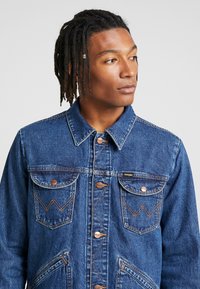 Jeansowa kurtka w ciemnym niebieskim kolorze z mosiężnymi guzikami, dwiema kieszeniami na piersi i wyraźnymi szwami. Posiada klasyczny kołnierz oraz teksturowany materiał.