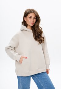 Beige hoodie met een zak aan de voorkant, geribde boorden en een relaxte pasvorm. Gecombineerd met lichtblauwe jeans. Glad textuur.
