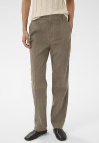 Persoon die taupe suède broek met voornaad draagt, beige geribd top ingestopt, en zwarte instapschoenen, staand met handen in zakken.