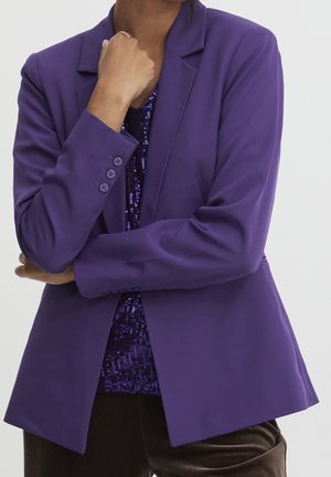 Personne portant un blazer violet sur un haut bleu foncé à sequins, une main levée près du menton et l'autre bras croisé.