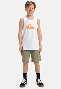 Witte tanktop met een oranje logo, gecombineerd met olijfgroene shorts. Zwarte sneakers met witte veters en zwarte crew sokken.