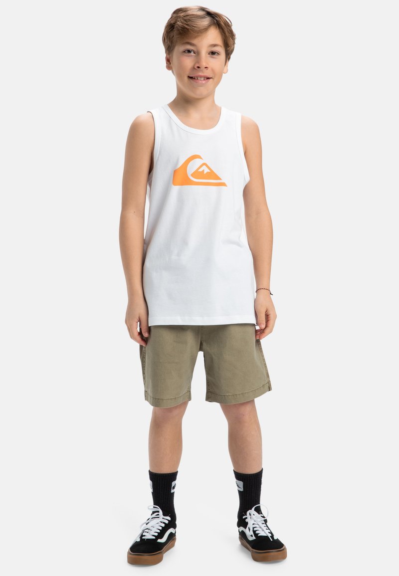 Witte tanktop met een oranje logo, gecombineerd met olijfgroene shorts. Zwarte sneakers met witte veters en zwarte crew sokken.