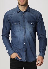 Tummansininen farkkupaita, jossa on napillinen etuosa, kaksi rintataskua läpillä ja kontrastivärinen ompelu. Pitkät hihat.