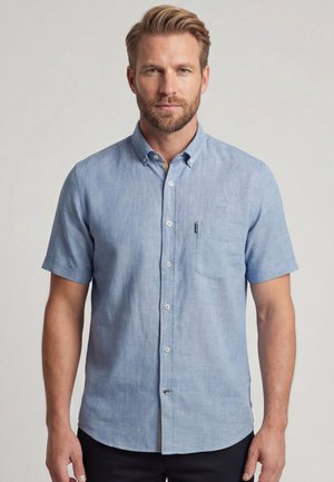 Mann mit Bart, der ein hellblaues kurzärmeliges Hemd mit Knöpfen trägt und vor einem schlichten Hintergrund steht, blickt geradeaus.