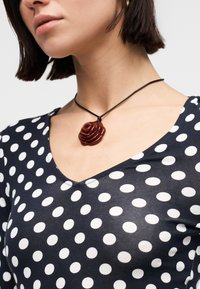 Γυναίκα που φοράει τοπ με μαύρα και λευκά πουά και μαύρο κορδόνι με μενταγιόν σε σχήμα σκούρου κόκκινου τριαντάφυλλου.