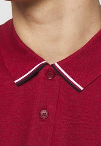 Czerwona koszulka polo o teksturowanym wyglądzie, z kontrastującym wąskim kołnierzykiem w czarne, białe i czerwone paski oraz okrągłym guzikiem z logotypem.