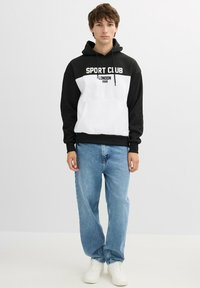 Sudadera en blanco y negro con el texto "SPORT CLUB LONDON 2028", con capucha ajustable y bolsillo canguro, combinada con jeans de color azul claro.