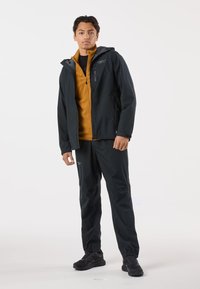 Herrens outdoor-outfit bestående av en svart vattentät jacka med huva och fickor med dragkedja, en senapsgul fleece under, och matchande byxor.