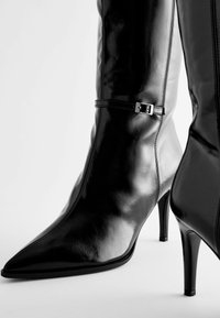 Juodų odinių iki kelių botų su smailiu nosiu, blizgiu paviršiumi ir metaline sagtimi ant kulkšnies. Stiletto tipo aukštakulniai.