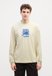 Beige hosszú ujjú póló, kerek nyakkal, kék grafikával, amelyen egy oroszlán és a "PR" valamint "HUGO" feliratok szerepelnek. Puha anyag, sima textúrával.
