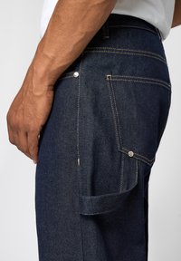 Mørkeblå denim jeans med gul syning, der har en baglomme, metalnitter og en sideløkke til at hænge ting op.