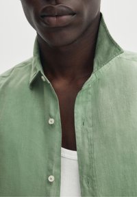 Grön linneskjorta med spetsig krage, knäppslå och texturerat tyg; lager-på-lager över en vit ribbad linne-topp.
