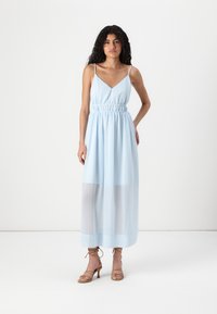 Ανοιχτού μπλε αμάνικο maxi φόρεμα με V λαιμόκοψη και συγκεντρωμένη μέση. Διάφανο ύφασμα καλύπτει το κάτω μισό. Το μοντέλο φοράει μπεζ ψηλοτάκουνες γόβες.