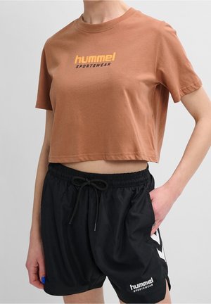 Persona vistiendo una camiseta corta marrón y unos pantalones cortos negros con cordón, ambos con el logo "hummel SPORTSWEAR", de pie con una mano en el bolsillo.