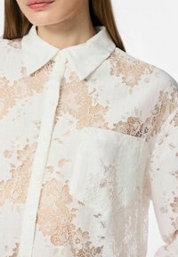 Witte kanten blouse met een kraag, knoopsluiting en een enkel zakje aan de voorkant. De stof heeft bloemmotieven en een doorzichtig effect.