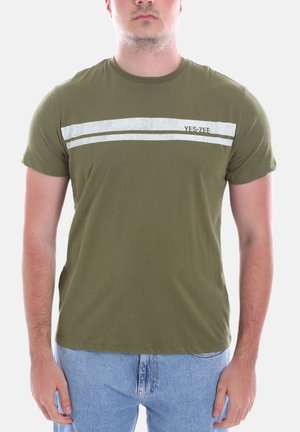 Uomo che indossa una maglietta verde oliva con due strisce orizzontali argento e testo "YES • ZFE", abbinata a jeans blu chiaro, in piedi davanti a uno sfondo bianco.