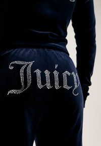 Tumšzilas velūra sporta bikses ar rhinestone rotājumiem, kas vārda "Juicy" uzraksts ir izvietots pa muguru, ar maigām tekstūrām un pieguļošu formu.