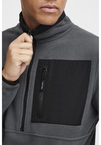 Pullover in pile grigio con un pannello nero e tasca a zip sul petto. Collo alto, tessuto testurizzato e dettagli a contrasto.